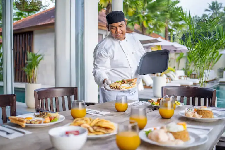 A person in a chef’s uniform delivers plates of food to a wooden table in a tropical poolside setting.