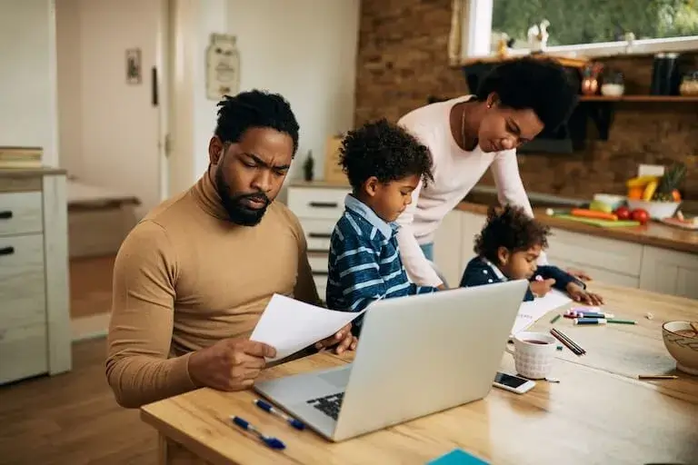 A person sits in a kitchen in front of a laptop and holds up a piece of paper next to another person helping two children with their homework