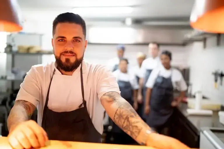 A person with a beard and tattoos wears a chef uniform and stands in a restaurant kitchen with several other people standing in the background.