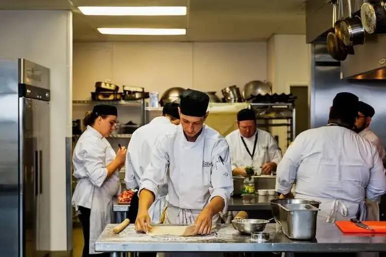 Three people in chef’s uniforms and hair coverings work side-by-side at a metal counter in a restaurant kitchen cutting vegetables, surrounded by bowls, containers, and other culinary items.