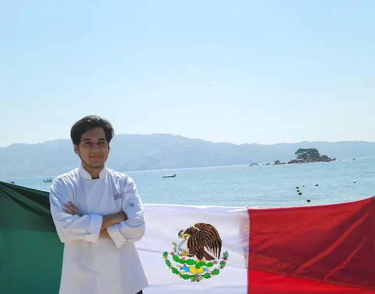 A young person in a chef’s uniform smiles and stands with arms crossed in front of a Mexican flag with a bay and mountains in the background.