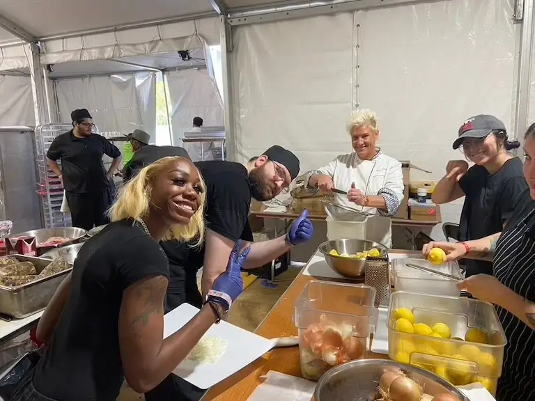 Culinary volunteers and Escoffier students prep ingredients at a long worktable inside a festival tent, smiling while chopping onions, zesting lemons, and straining citrus alongside chef Anne Burrell.