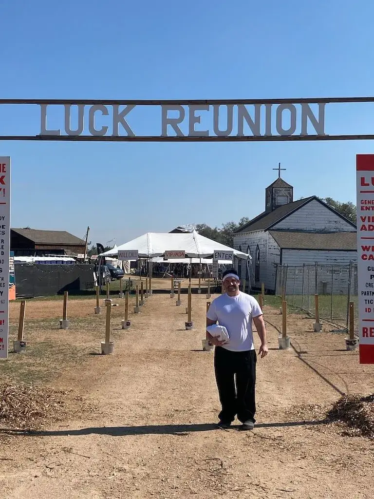 Escoffier graduate Michael Fields, wearing a white tee shirt and black pants, stands on a dirt road under a sign reading “Luck Reunion.”