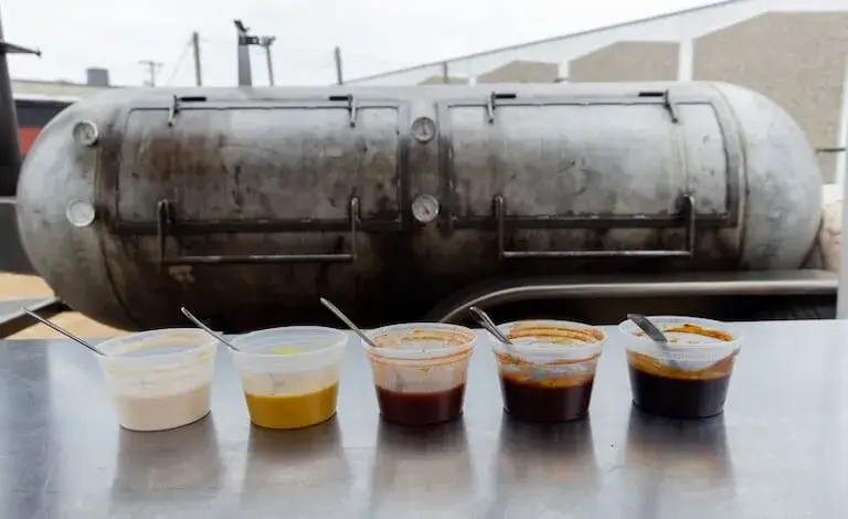 Five sample cups of different colored sauces ranging from light cream to dark brown, lined up in front of a large smoker for testing and development