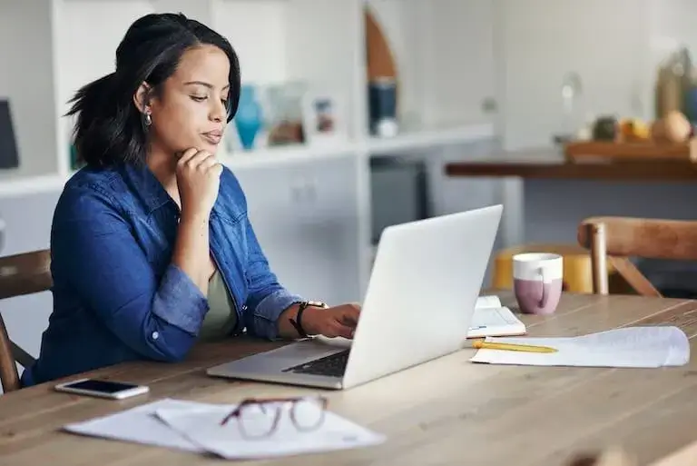 A person in a denim shirt sits at a wooden kitchen table and looks thoughtfully at a laptop surrounded by notebooks, papers, a cup of coffee, a smartphone, and a pair of glasses.
