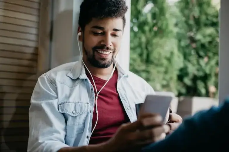 A smiling person with a beard and wearing wired earbuds sits in a bright window holding a mug of coffee and a smartphone.