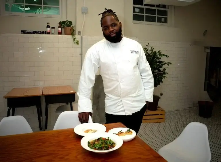 Chef Rodney Smith in white chef coat standing behind table with three plated dishes in an intimate dining space