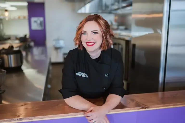 Chef Ellen Doerr wears her “Chef Ellen” branded chef’s coat while smiling in her professional kitchen.