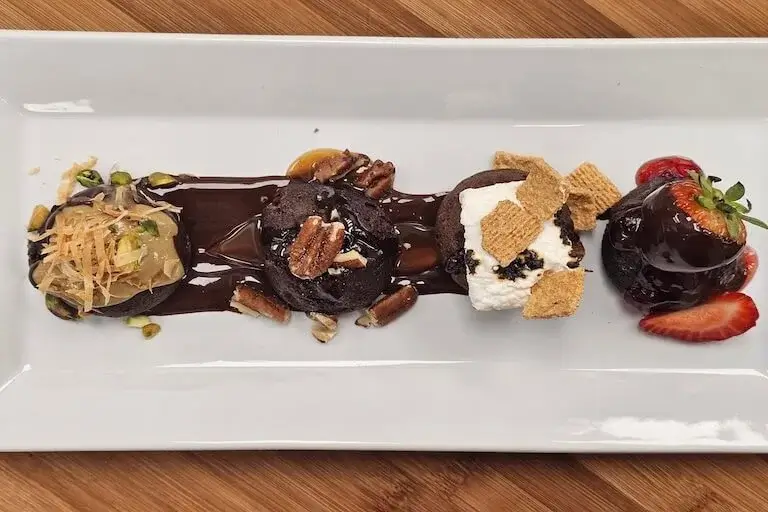 Artfully plated chocolate dessert flight featuring multiple components including chocolate bites, pistachios, pecans, toasted marshmallows, and fresh strawberries.