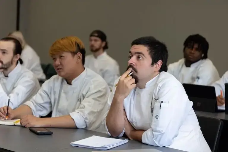 Several students wearing Escoffier-branded chef’s jackets sit at long tables in a classroom listening attentively.