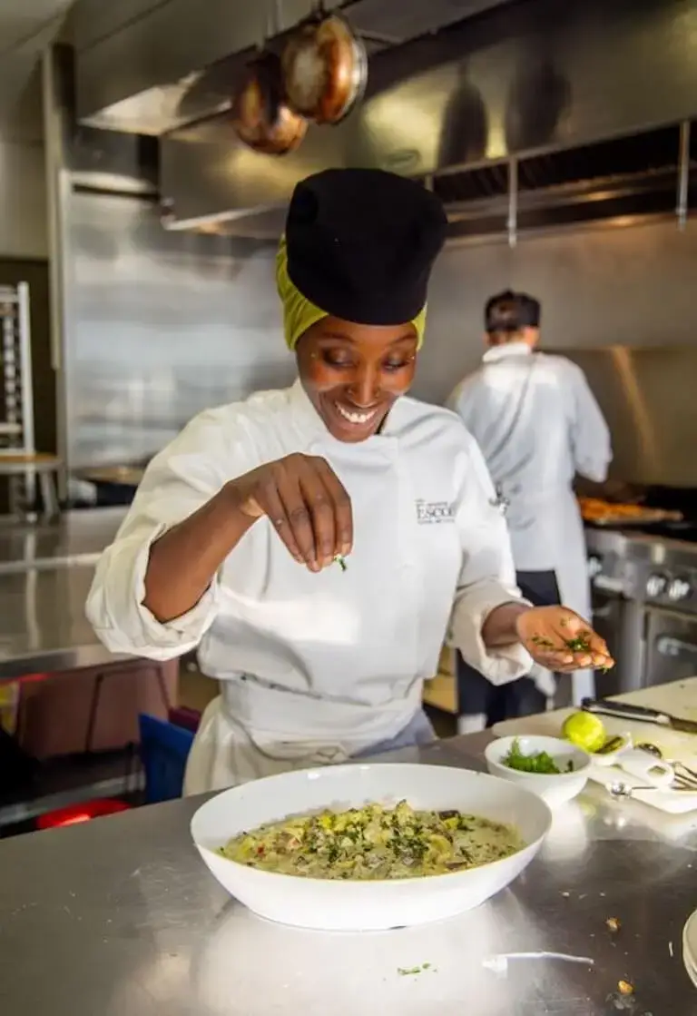 Culinary student sprinkling fresh herbs over a finished dish while plating in a professional kitchen.