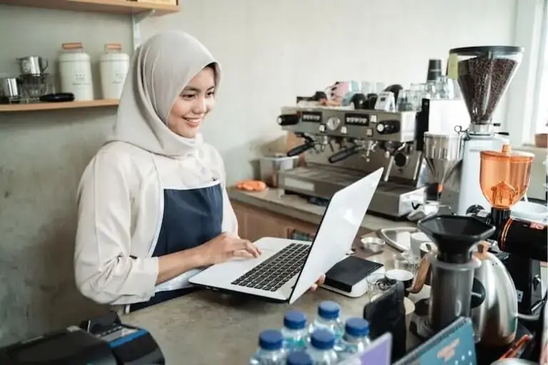 Café owner in apron working on laptop at coffee bar with espresso machine in background.