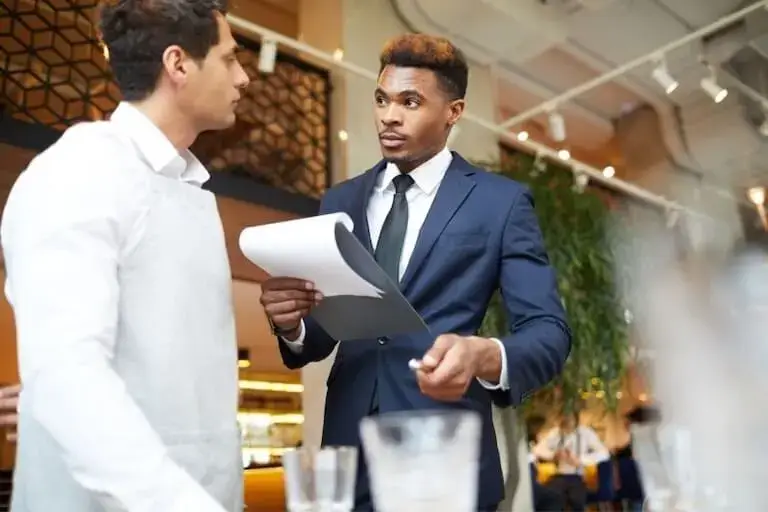 A manager holding a clipboard speaks with a staff member in a restaurant dining room.