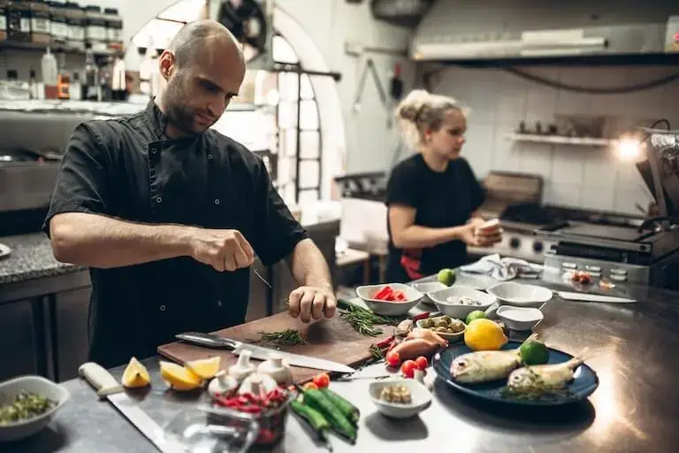 A person in a black chef’s uniform strips rosemary leaves from the stem at a stainless steel counter covered with other ingredients in a restaurant kitchen while another person in a black short-sleeved shirt walks past.