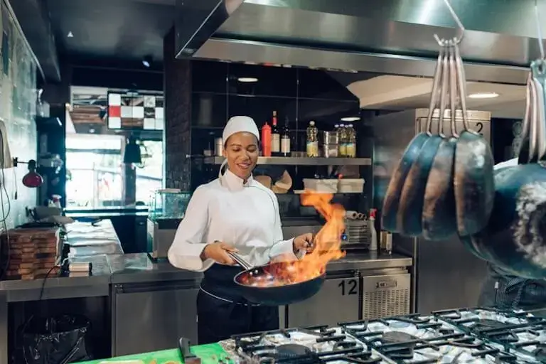 A person in a white chef’s uniform and cap holds a flaming sauté pan in a restaurant kitchen.