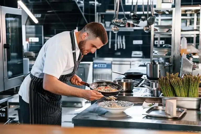 A person in a chef’s uniform carefully transfers pasta from a skillet to a serving dish in a restaurant kitchen.