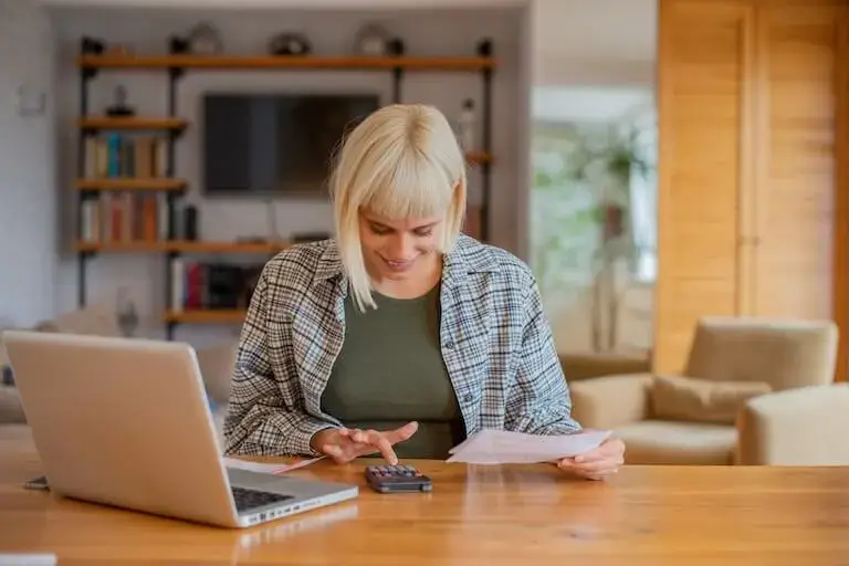 A person with blond hair and wearing a checkered shirt smiles as they use a calculator and hold a piece of paper next to an open laptop in a modern living room.