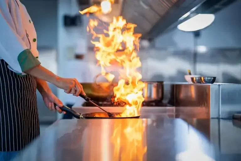 Chef sautéing food in a pan with tall flames rising on a stovetop.