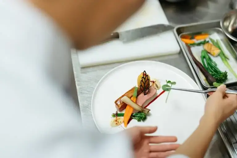 A chef using tweezers to place garnishes on a plated dish in a professional kitchen.
