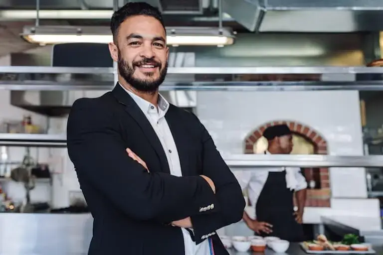 A person with a beard and wearing a dark blazer smiles and stands with arms crossed in a modern restaurant kitchen while a person in a chef’s uniform stands in the background.