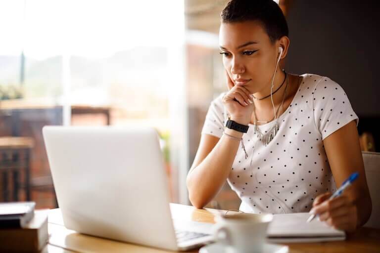 A person with short hair and wearing a short-sleeve polka-dot t-shirt listens to music with wired earbuds while looking at a laptop and writing something in a notebook while seated in a café.