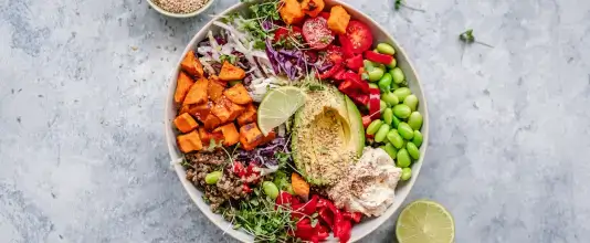 Buddha bowl with roasted sweet potatoes, sliced avocado, quinoa, edamame, shredded cabbage, sprouts, and colorful vegetables arranged in a bowl on a light surface.