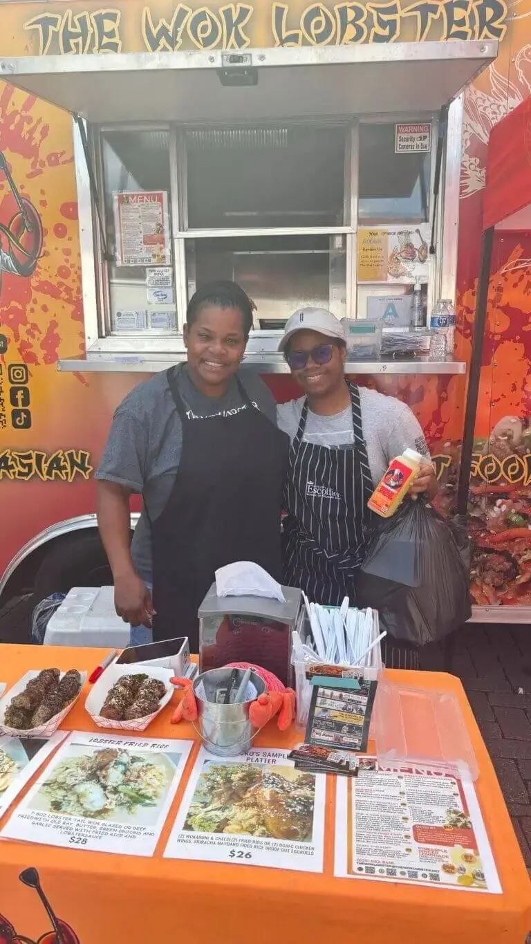 Two smiling people in aprons stand in front of a bright orange "The Wok Lobster" food truck. In the foreground, a table displays menu signs for lobster fried rice and sample platters alongside baskets of prepared food.