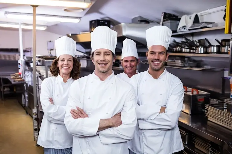 Four chefs wearing white chef coats and tall toques stand side by side in a commercial kitchen, smiling with their arms crossed. Stainless steel counters, shelves of cookware, and kitchen equipment are visible in the background.