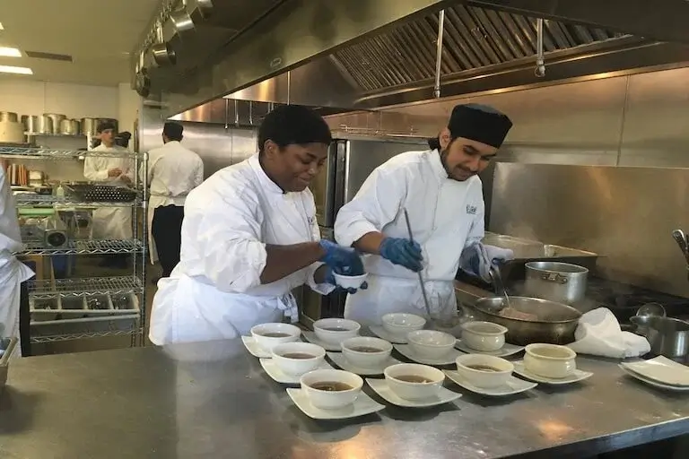 Two culinary students in white chef coats plating soup dishes in a professional kitchen, with additional students working in the background