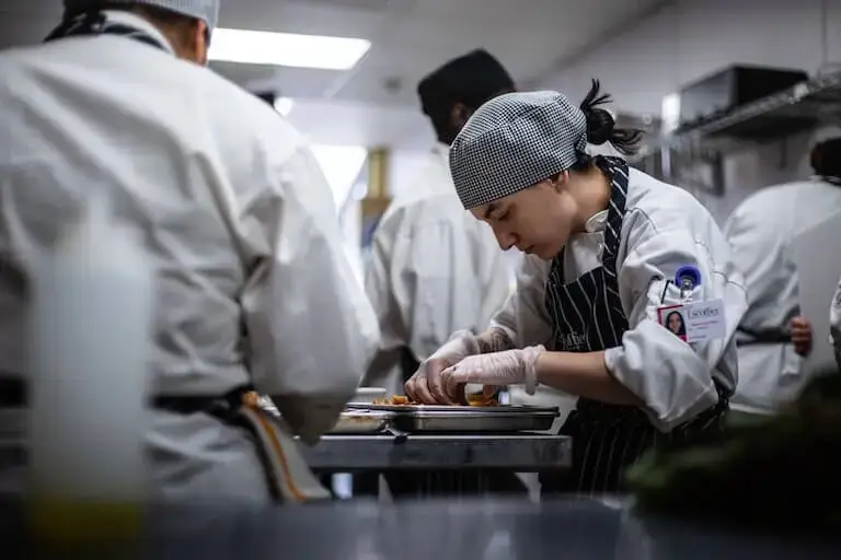 A person wearing a chef’s uniform, a checkered cap, and disposable gloves carefully arranges ingredients on a baking sheet in a busy professional-grade kitchen.