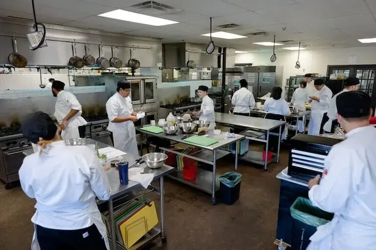 Several people wearing chef’s uniforms and black caps prepare ingredients, read instructions, and perform other culinary tasks in a professional-grade kitchen.