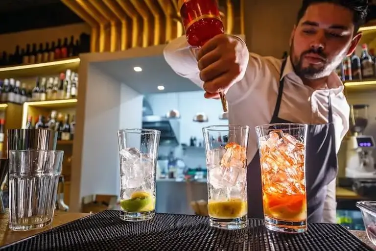 A bartender in an apron carefully pours ingredients into cocktail glasses at a bar with bottles visible in the background.