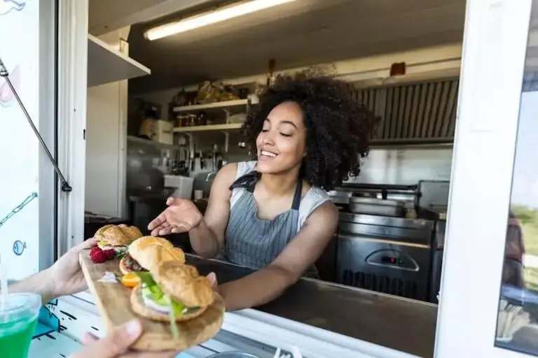 Smiling woman in blue apron cooking sandwich in home kitchen with laptop open nearby