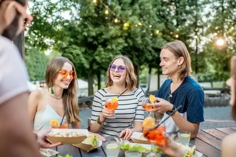 Three friends laughing and enjoying orange drinks together at an outdoor cafe table with food.