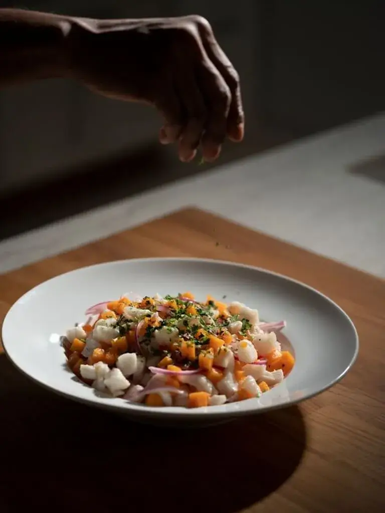 Hand sprinkling chopped fresh herbs over a plated dish of diced vegetables and seafood on a wooden countertop.