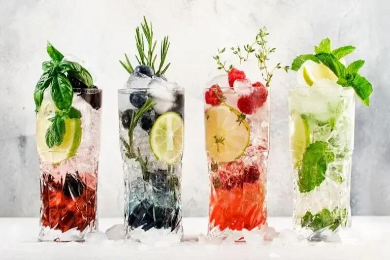 Four colorful non-alcoholic mocktails garnished with fresh herbs and fruit arranged on a white marble surface.