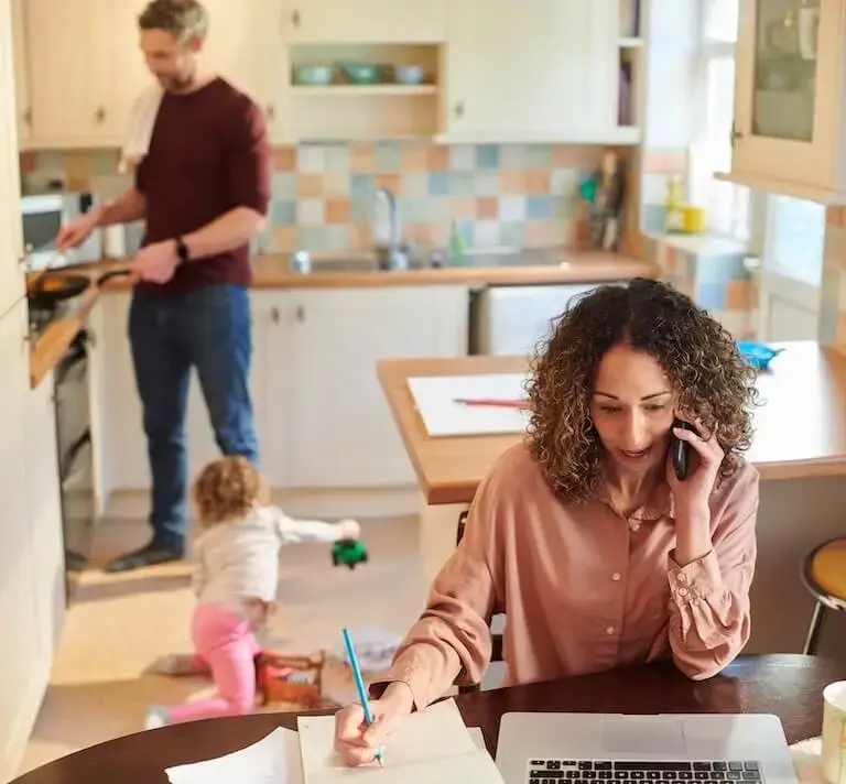 A person with long curly hair speaks on a cell phone while writing in a notebook and looking at a laptop in a home kitchen while in the background another person cooks at the stovetop and a child plays with toys on the floor.