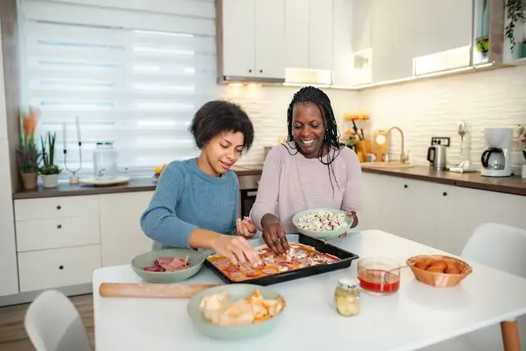 A close-up photograph of an adult and a teenager smiling while making homemade pizza together in a kitchen. The teenager is focused on placing toppings onto the dough, while the adult stands beside them holding a bowl of ingredients.