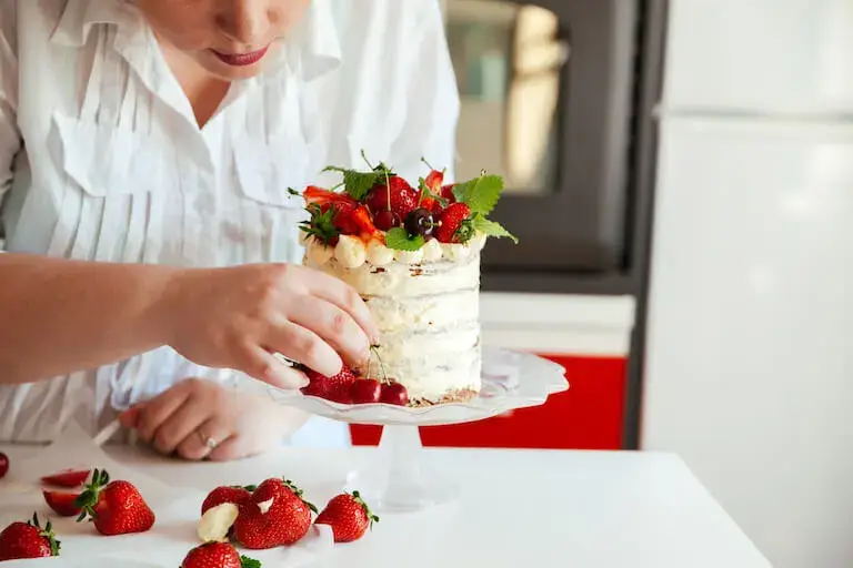 Close-up of a baker in a white shirt garnishing a small cake with strawberries, cherries, and mint leaves. Fresh strawberries and whipped cream are scattered on the white tabletop in the foreground.