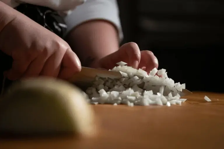 Close-up of a person using a chef’s knife to create a uniform small dice of white onion on a wooden cutting board.