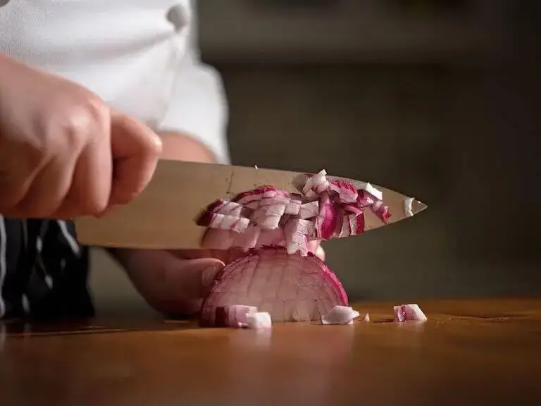 Closeup of chef’s hands dicing a red onion with a chef’s knife on a wooden cutting board, with small diced pieces scattered nearby.