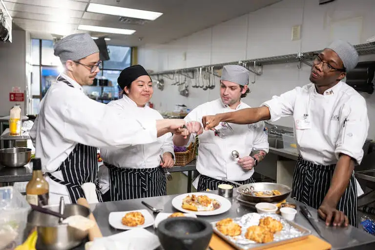 Culinary students celebrating with a fist bump in a commercial kitchen surrounded by prepared dishes.