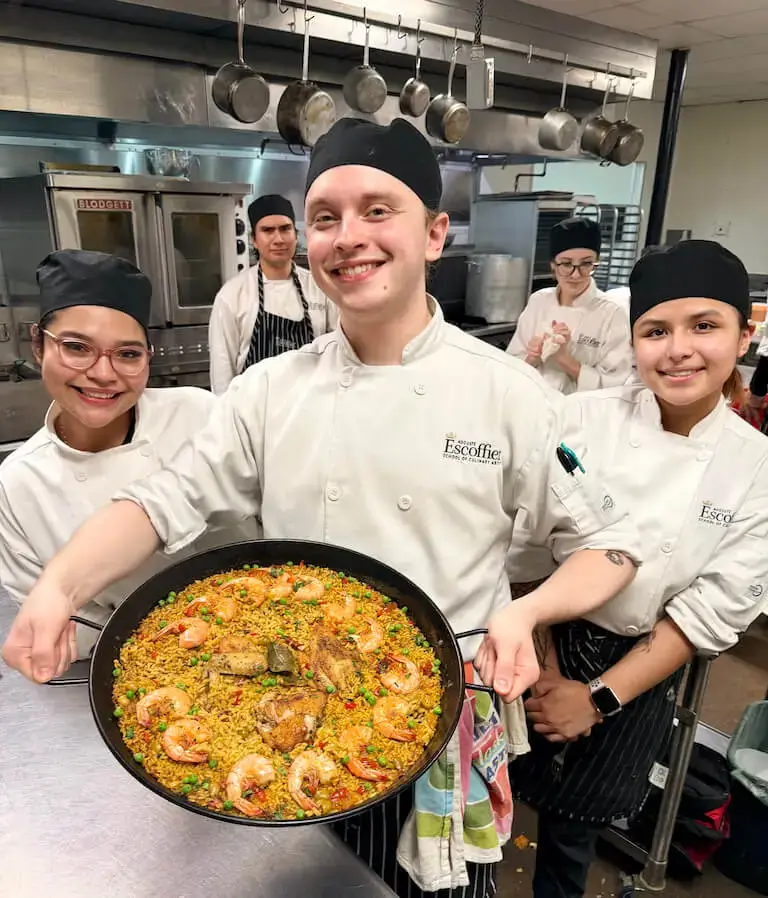 Three Escoffier culinary students presenting a completed paella dish in a professional training kitchen with stainless steel equipment and hanging pots visible in background.