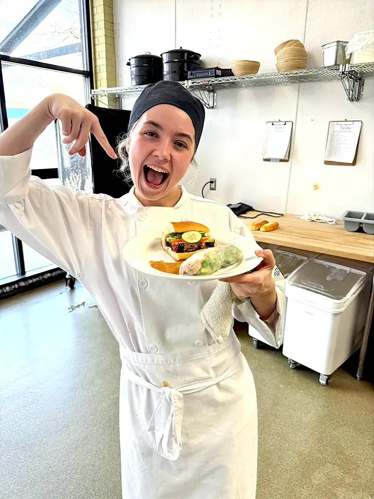 Excited culinary student in chef uniform presenting a plated sandwich with garnish and sides in a training kitchen.