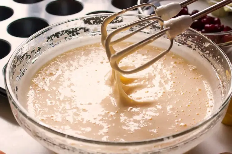 Batter dripping from mixer attachments into a clear bowl with more batter.