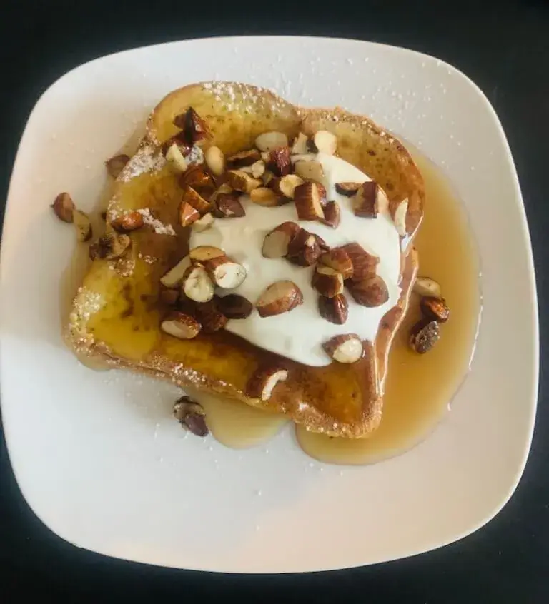 French toast topped with whipped cream, chopped hazelnuts, powdered sugar, and syrup on a white square plate.