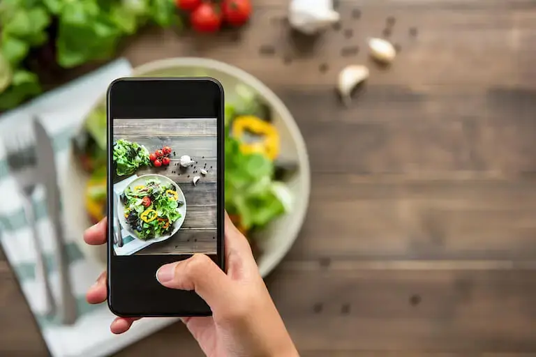 Hand holding smartphone photographing a colorful plated salad with fresh vegetables on a wooden table with blurred ingredients in the background.