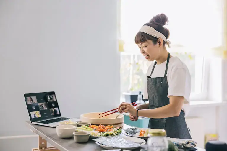 Person in apron teaching an online cooking class via laptop video call, preparing Asian cuisine with fresh ingredients including vegetables and sushi on a white counter in a bright kitchen