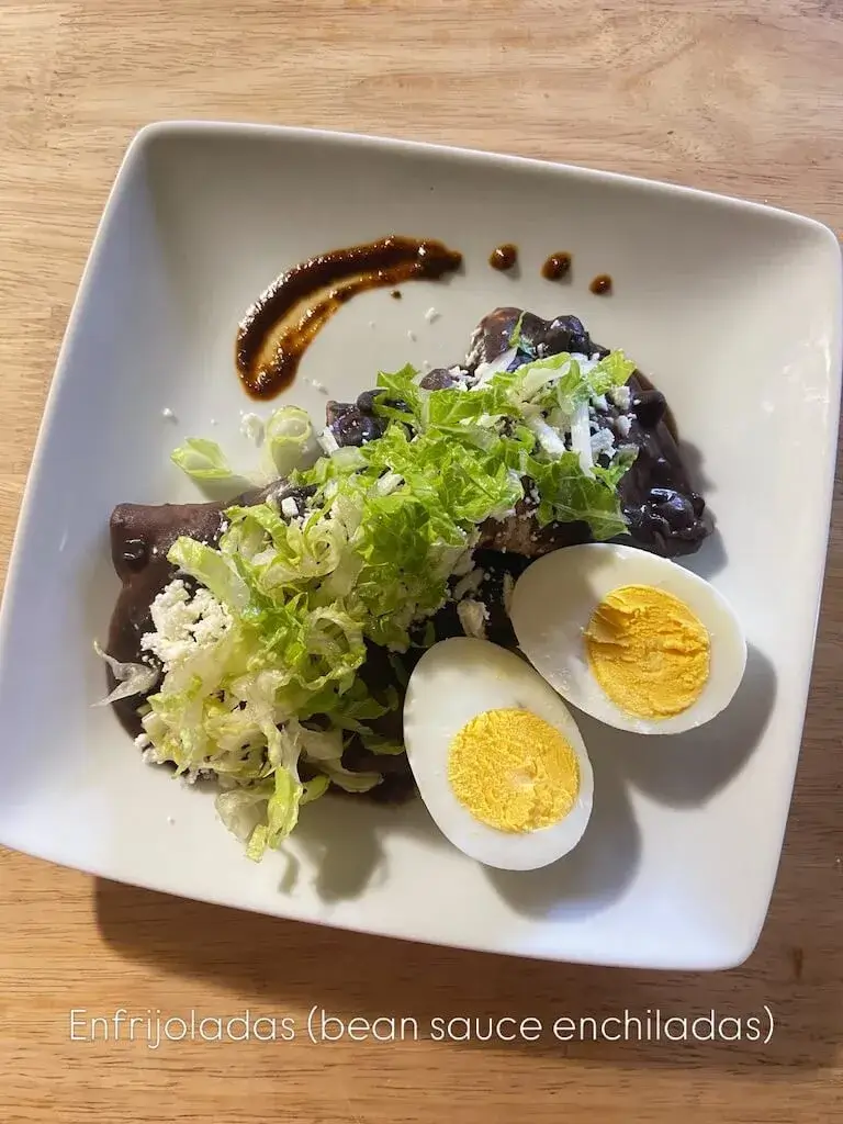 Plated enfrijoladas with sliced hard-boiled eggs, fresh greens, and bean sauce on a white rectangular plate with wooden background.