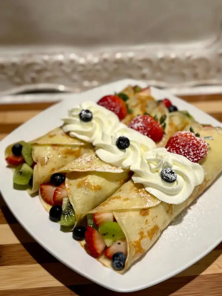 Three crepes filled with sliced strawberries, kiwi, and blueberries, topped with swirls of whipped cream and a dusting of powdered sugar on a white square plate.